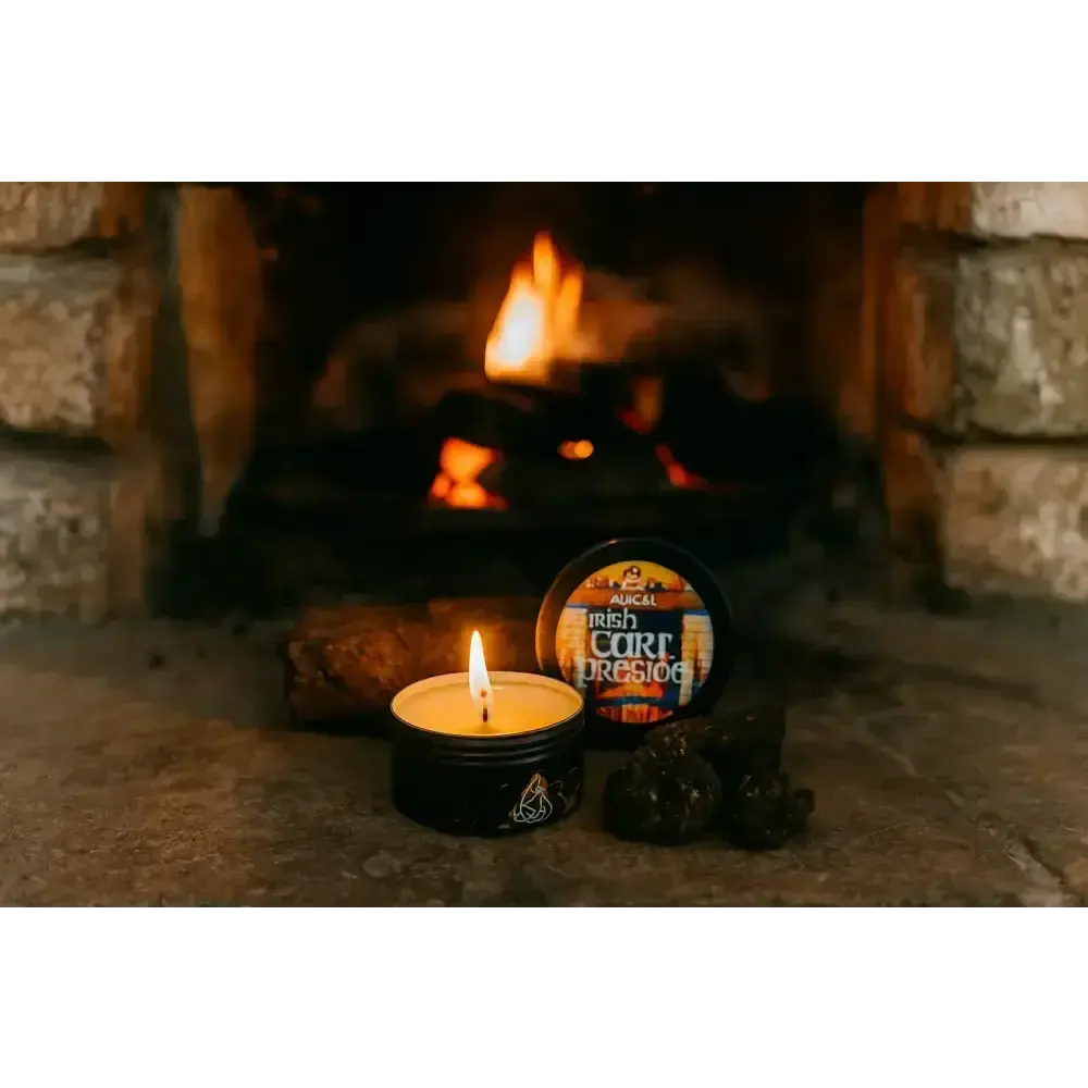 Lit Irish Turf Fireside tin candle with its lid and turf pieces on a hearth, a fire burning behind