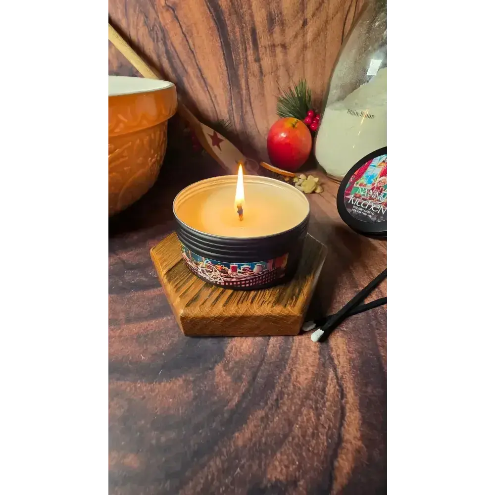 Lit Nanny's Kitchen tin candle on a wooden stand with a mixing bowl and apple behind